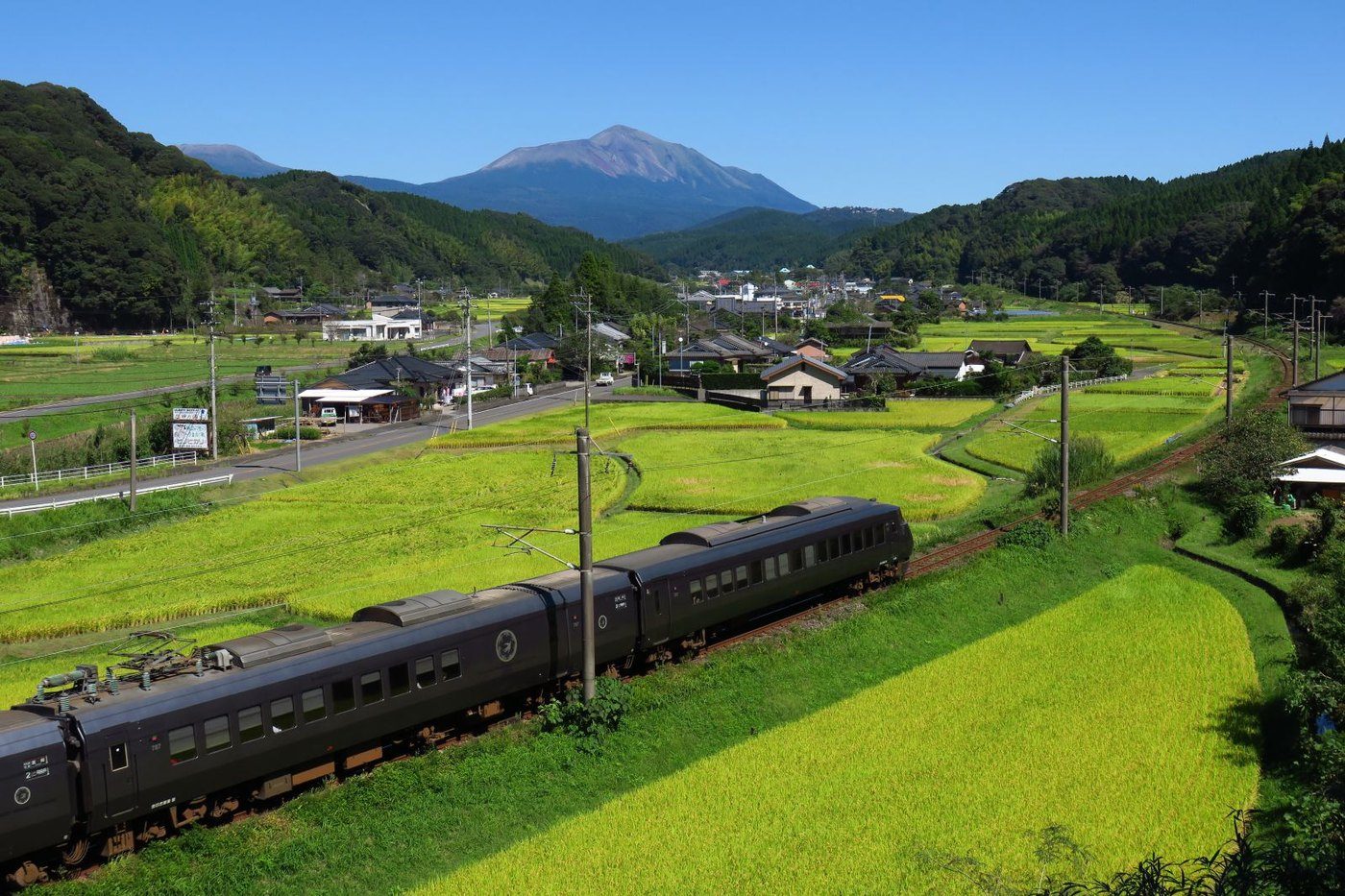 実りかけた田んぼの中を走る黒い列車。奥に山なみと集落が見えます。